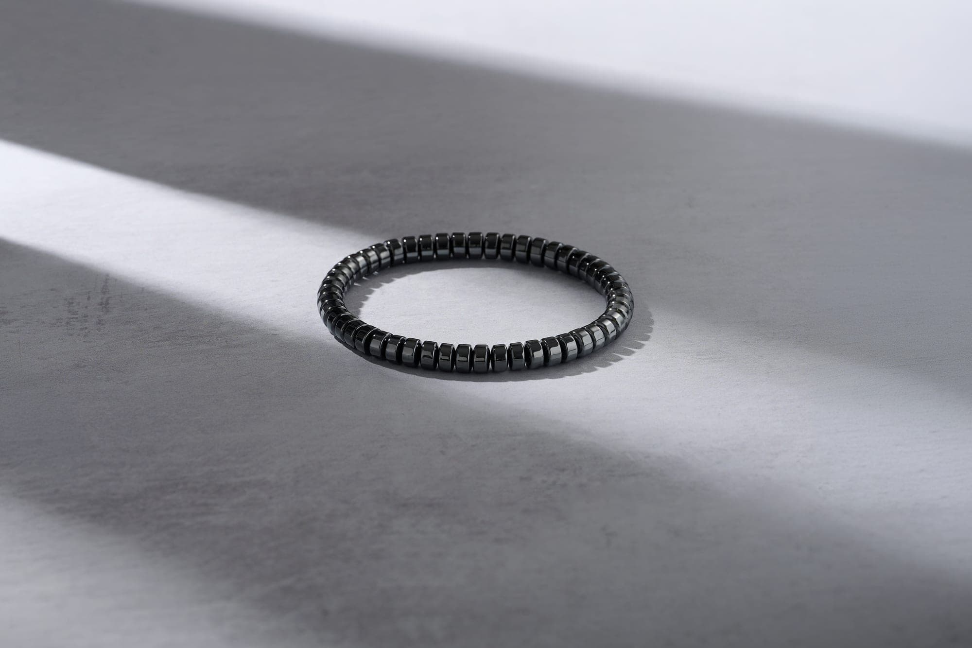 The IONISS hematite bracelet resting on a pale surface, casting a hard diagonal shadow in raking light